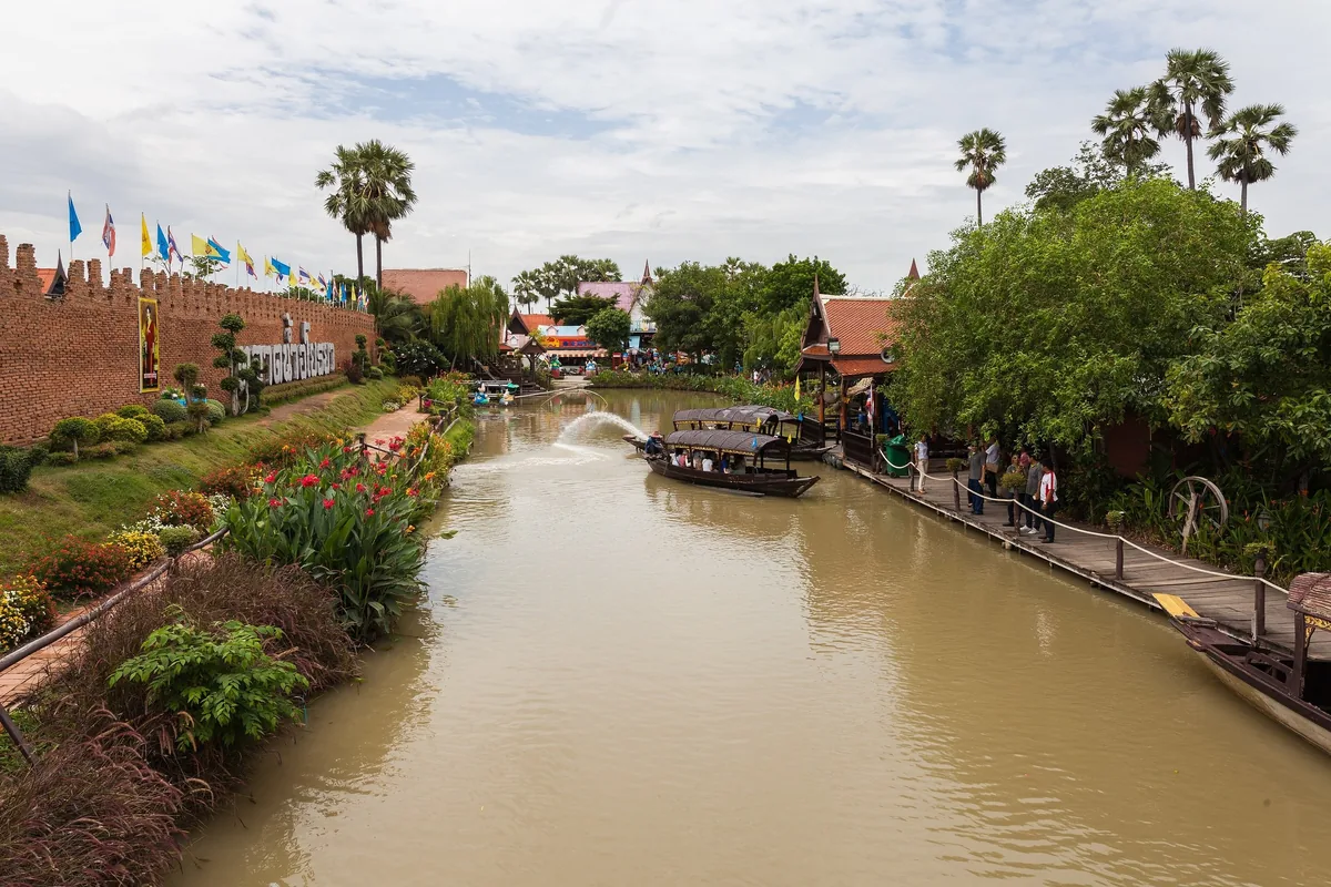 A tour boat glides along a canal past a fortress-style wall and wooden walkways at Ayutthaya's floating market.