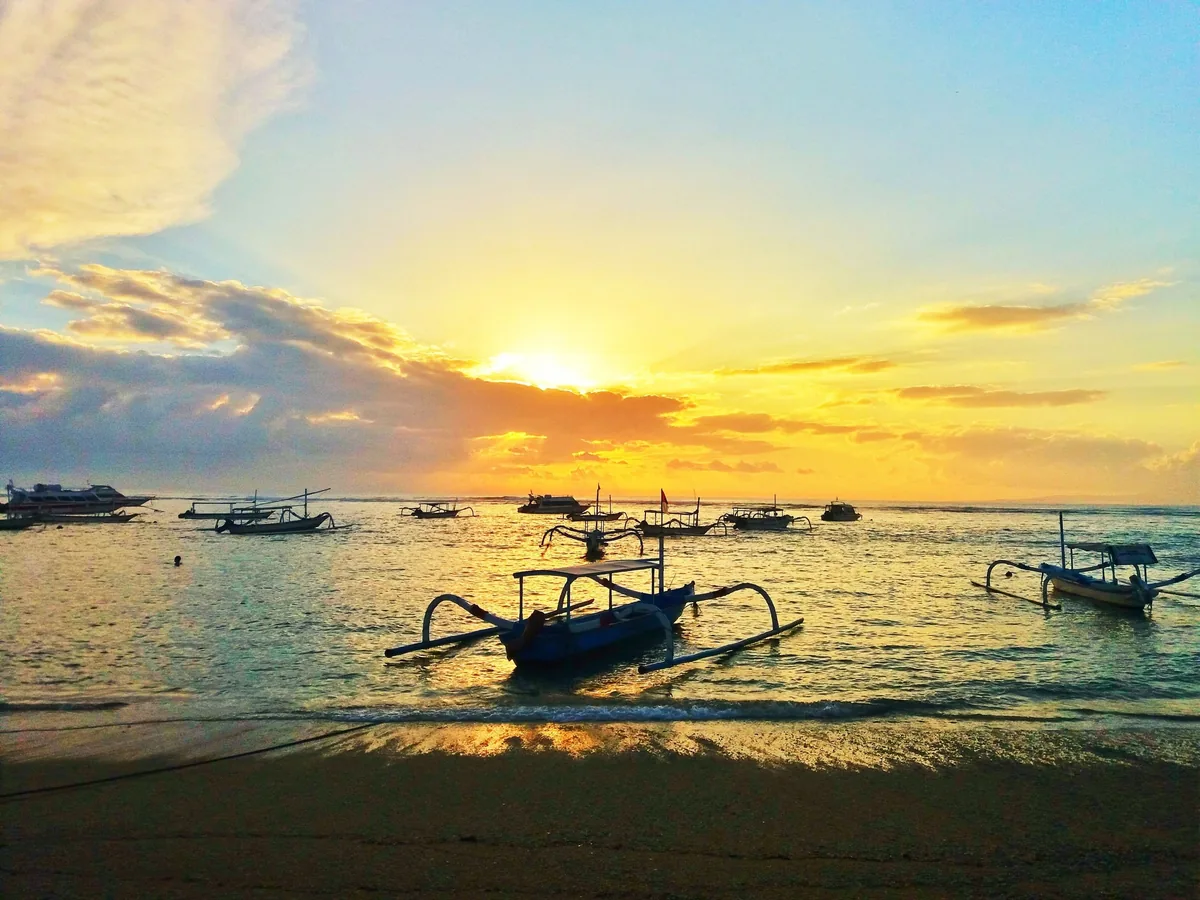 Traditional outrigger boats float on calm water at sunrise on Sanur Beach, Bali.