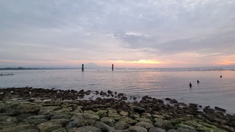 Sunset over calm sea with rocky shore, harbor markers, and people wading at Sanur beach in Bali