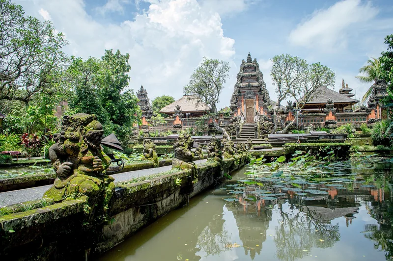 Moss-covered stone statues line a lotus pond leading to the tiered pagodas of Ubud Water Palace in Bali.