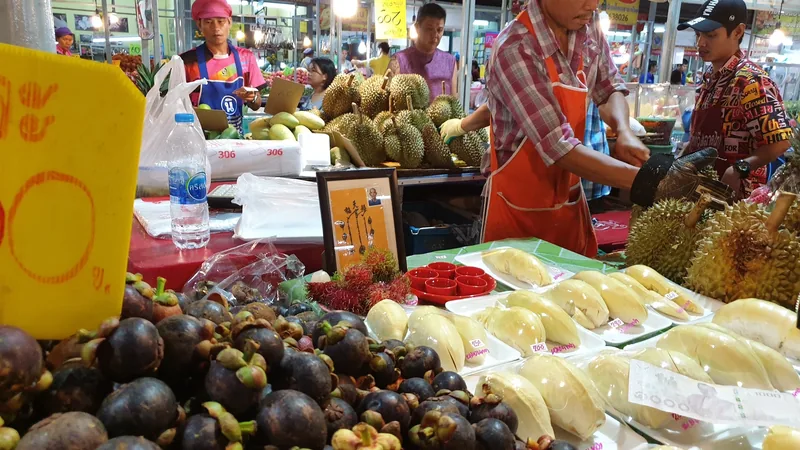 A market stall in Bangkok displays mangosteen, rambutan, and peeled durian while a vendor prepares fruit for sale.