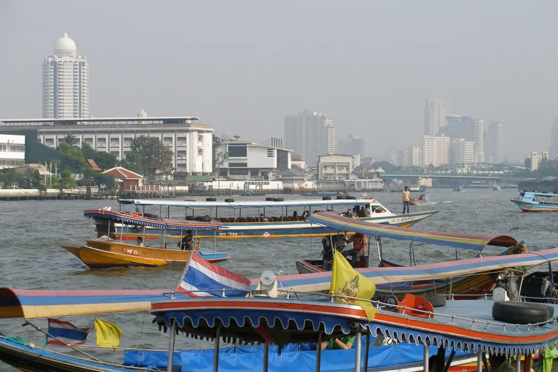 Long-tail boats with Thai flags crowd the Chao Phraya River beneath Bangkok's hazy skyline.