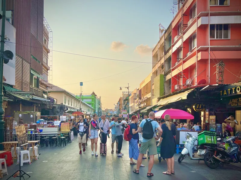 Tourists stroll along Khao San Road at sunset, lined with shops, tattoo parlors, and street vendors in Bangkok.