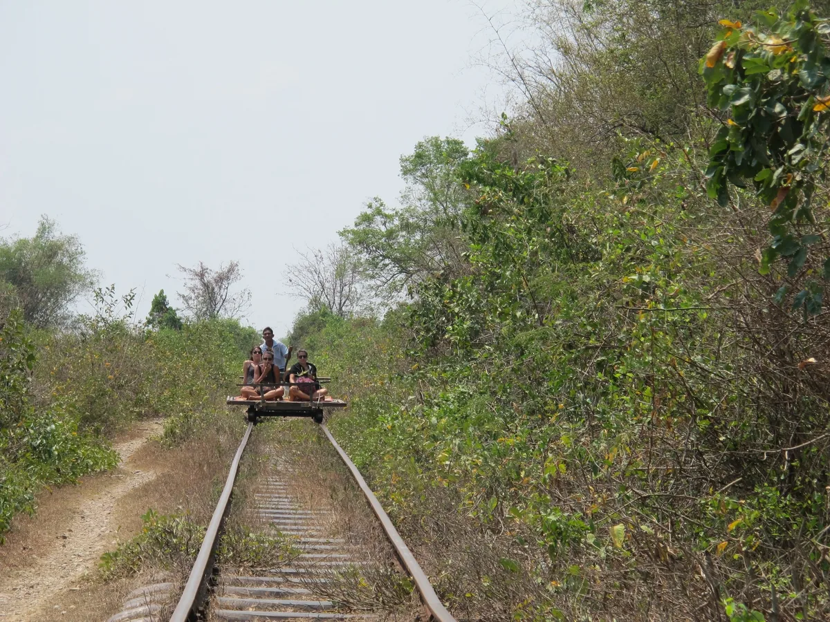 Tourists ride a bamboo train platform along a rural rail track lined with dense bush near Battambang.