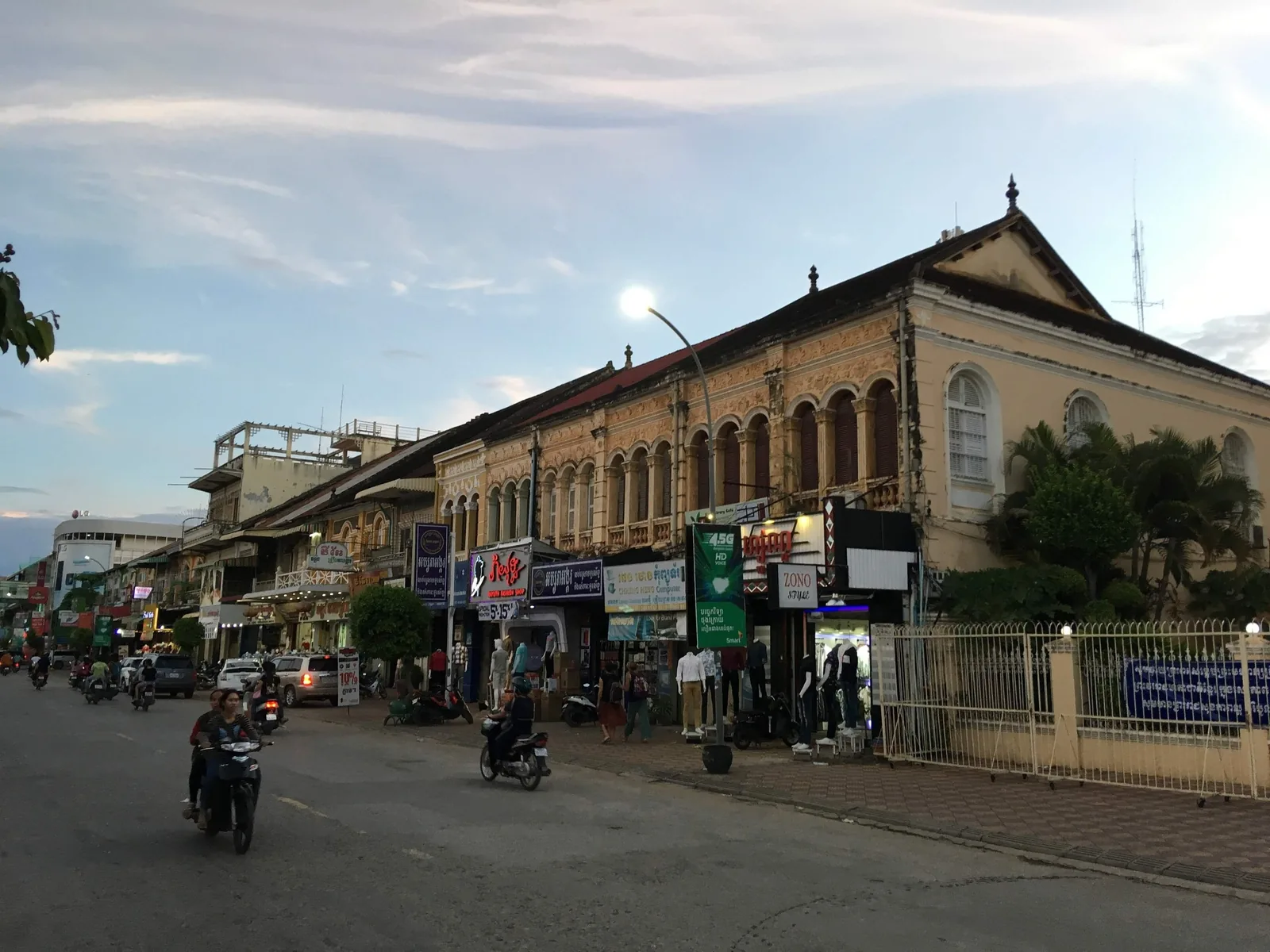 Colonial-era shophouses line a street at dusk in Battambang, with motorbikes passing by.