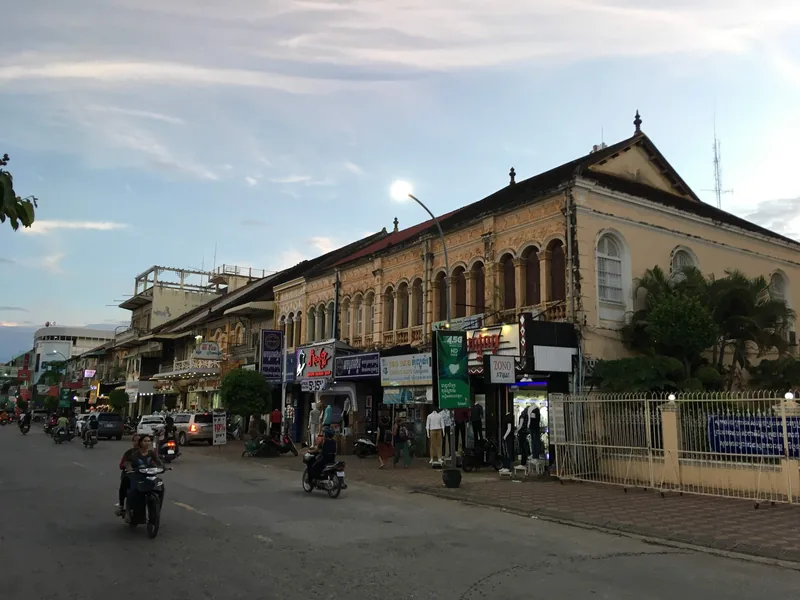 Colonial-era shophouses line a street at dusk in Battambang, with motorbikes passing by.