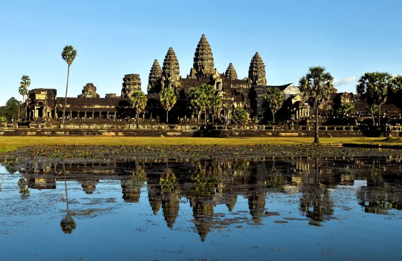 The towers of Angkor Wat rise above palm trees, reflected in a still pond under a clear sky in Siem Reap.
