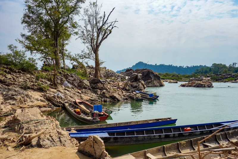 Wooden longtail boats moored along a rocky riverbank amid the Mekong islands of Si Phan Don, Laos.