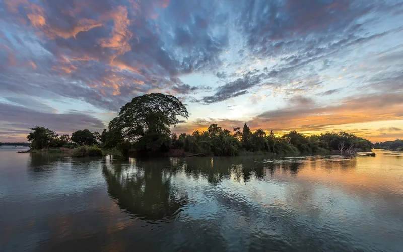 Colorful sunset clouds reflect over calm river waters around a tree-covered islet in Si Phan Don, Laos.