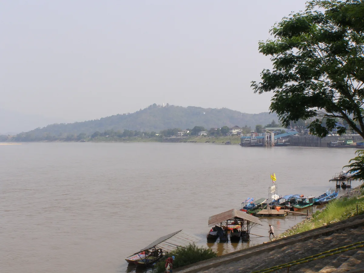 Wooden boats moored on the muddy Mekong River at Chiang Saen, Chiang Rai, with hazy hills beyond.
