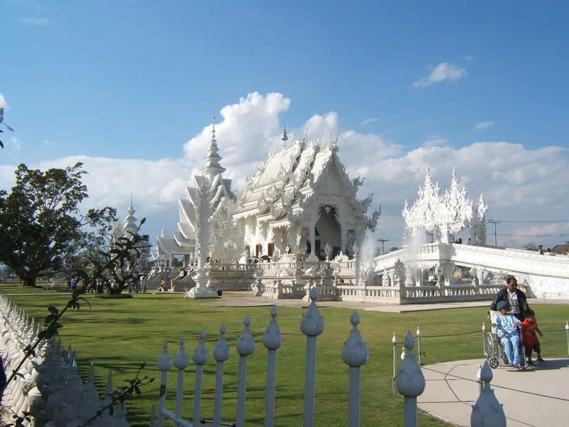 The intricate white spires of Wat Rong Khun rise above green lawns under a bright blue sky in Chiang Rai.