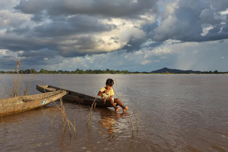 A boy sits on a wooden boat by the Mekong River in Si Phan Don under a dramatic cloudy sky.