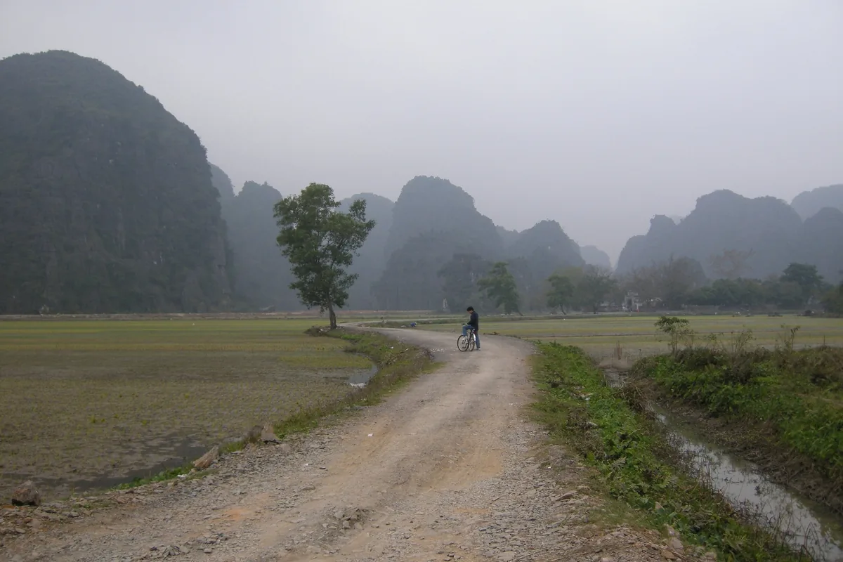 A cyclist pauses on a dirt path through rice fields beneath misty limestone karsts in Ninh Binh.