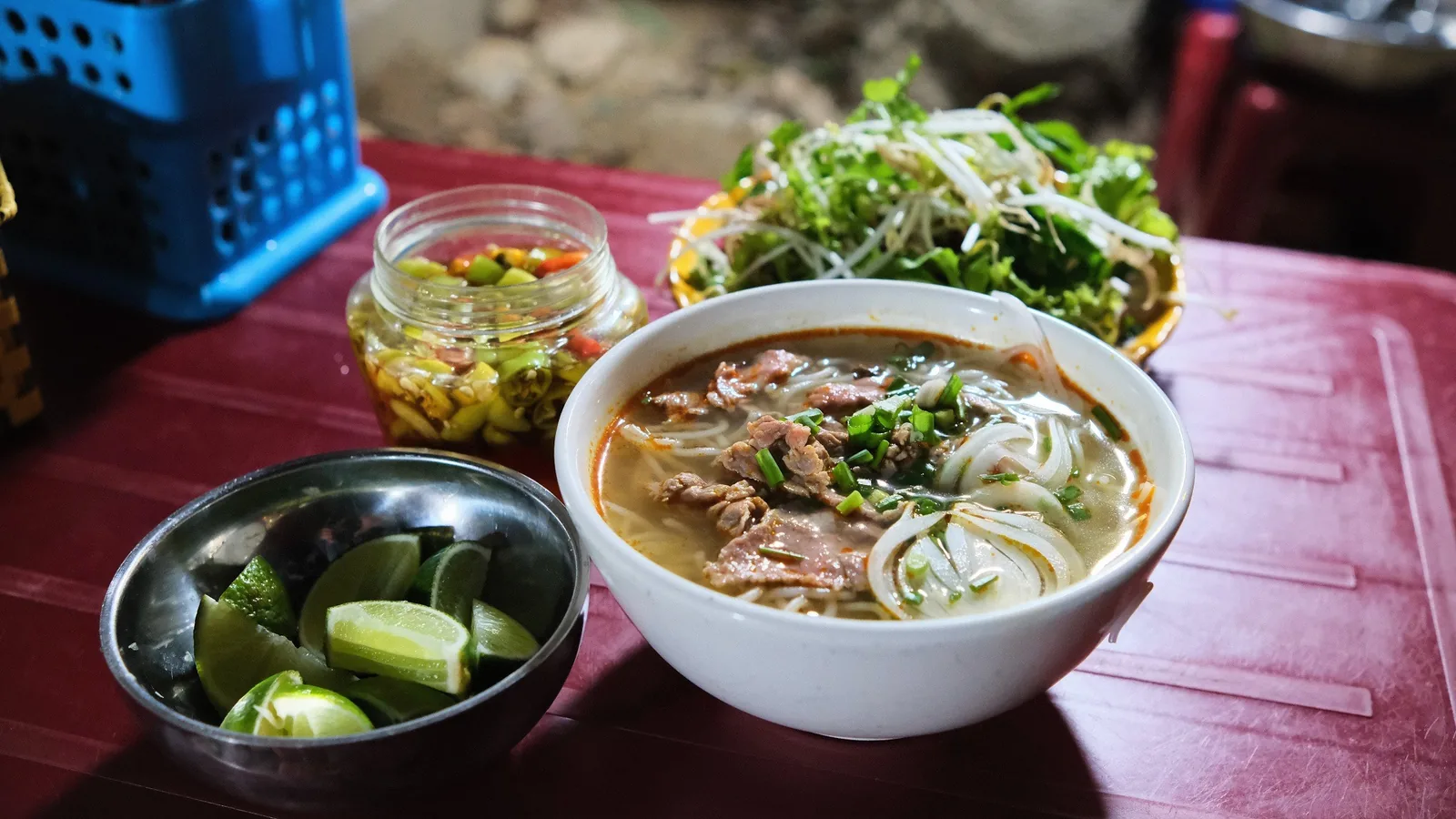 A bowl of Hue beef noodle soup served with lime wedges, pickled vegetables, and fresh herbs on a red table.