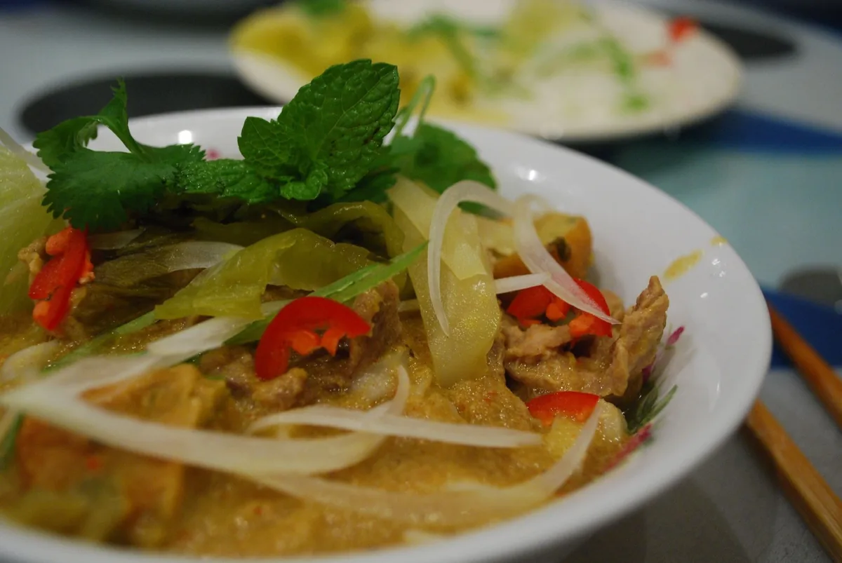 A close-up bowl of Thai khao soi curry topped with fresh herbs, chili, and pickled mustard greens.