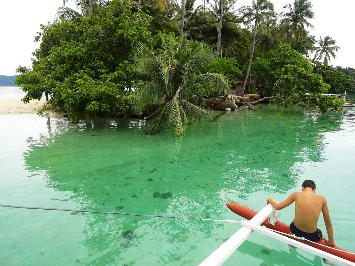 A man sits on an outrigger boat over turquoise water beside a small palm-covered islet near Port Barton, Palawan.