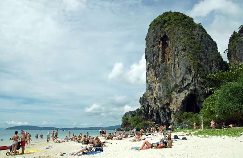Sunbathers relax on the white sand of Phra Nang Beach beneath a towering limestone cliff in Krabi.