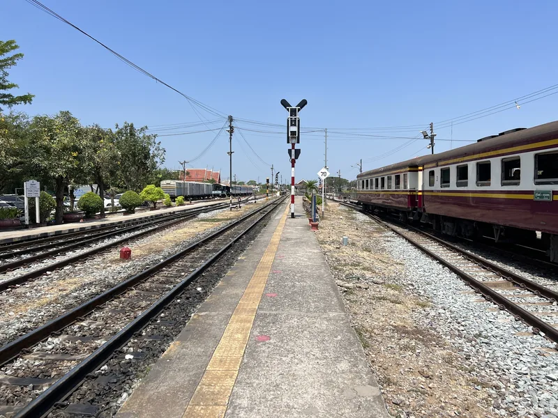 A maroon train carriage stands at a quiet platform lined with tracks and signal poles at Ayutthaya train station.