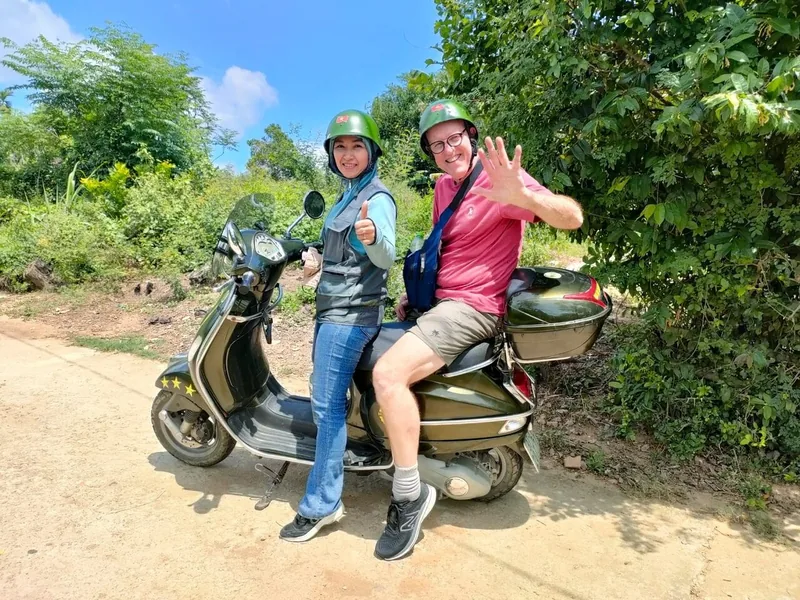 A tourist and local guide pose on a vintage Vespa scooter on a rural road near Hoi An.