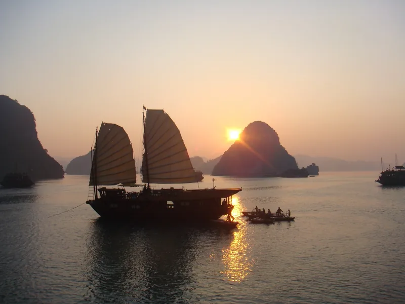 A traditional junk boat with sails silhouetted against the sunset among limestone karsts in Ha Long Bay.