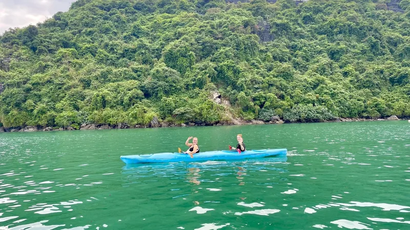 Two women paddle a blue kayak on emerald water beneath forested limestone cliffs in Lan Ha Bay.