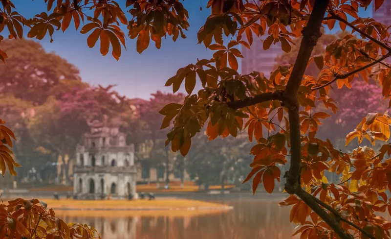 Turtle Tower stands on Hoan Kiem Lake in Hanoi, framed by autumn-colored tree branches.