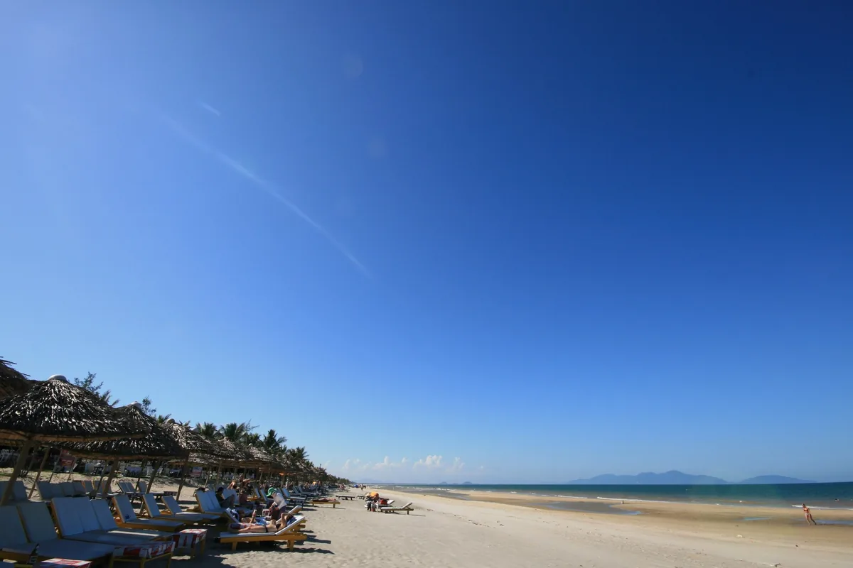 Thatched umbrellas and loungers line a wide sandy beach under a vast blue sky in Hoi An.
