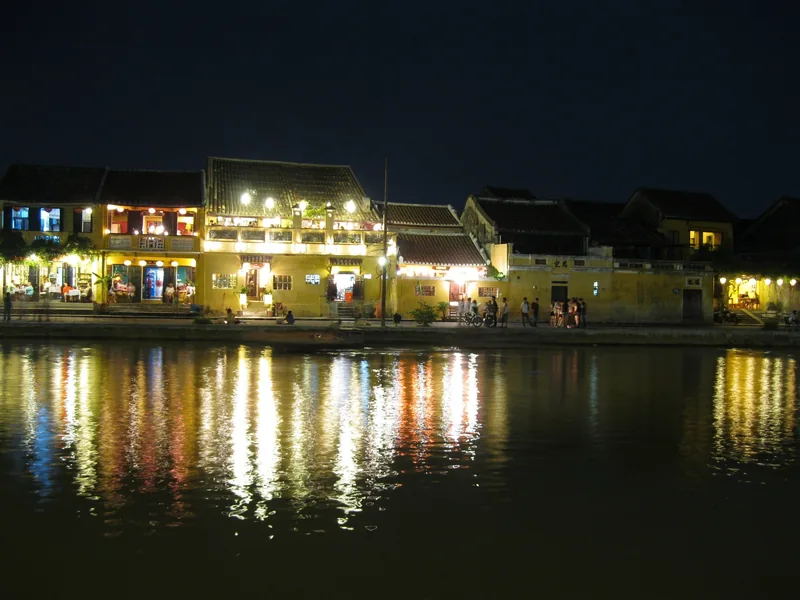 Illuminated old town buildings along the riverfront in Hoi An reflect on the water at night.