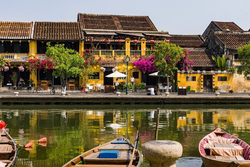 Yellow riverside houses with red lanterns and bougainvillea reflect in the calm river in Hoi An's old town.
