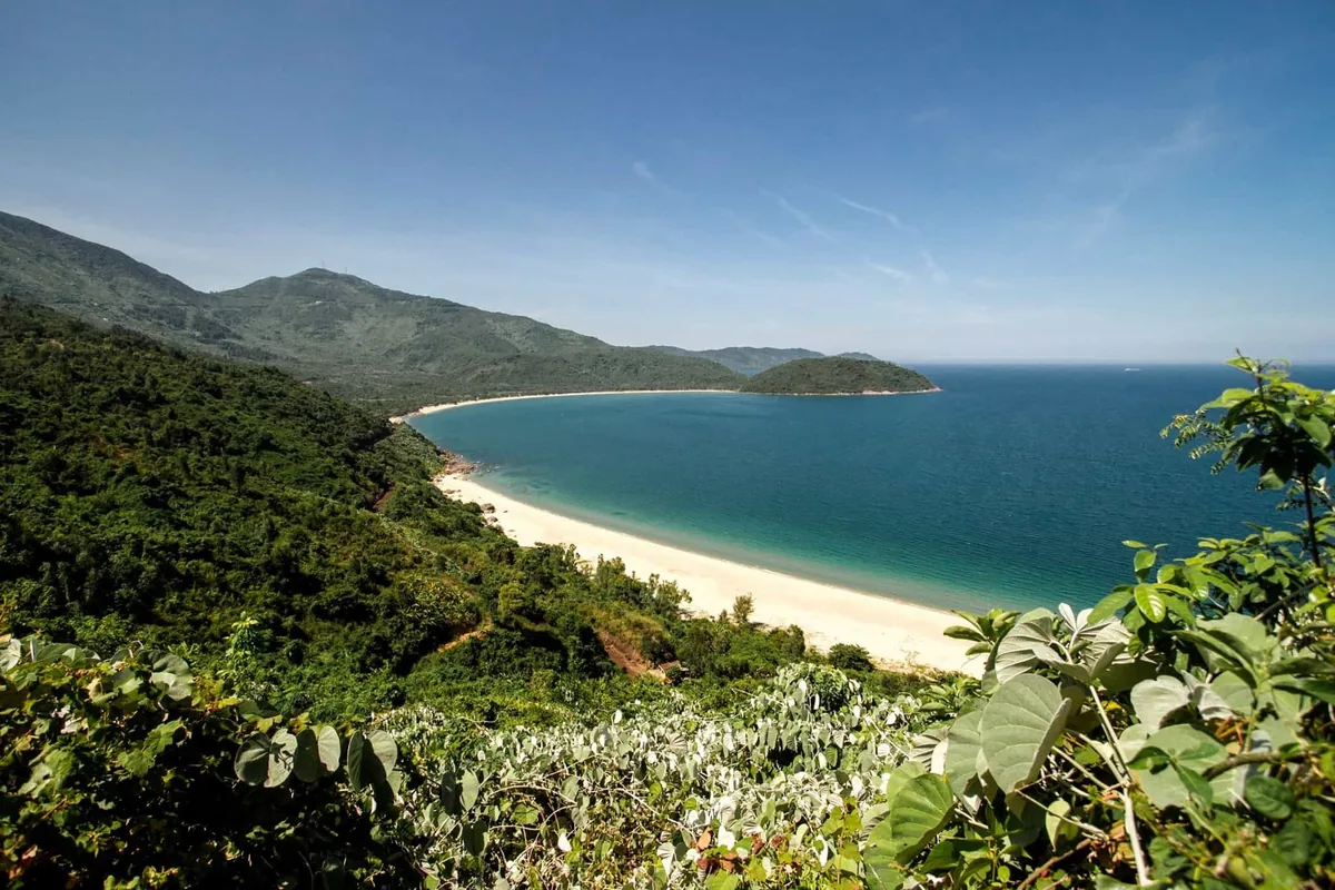 A curved white-sand beach and turquoise bay seen from a jungle-covered hillside near Hai Van Pass.