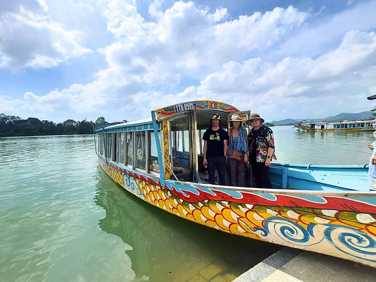Tourists stand aboard a colorful dragon boat on the Perfume River in Hue under a bright sky.