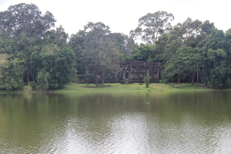 An ancient stone temple ruin sits among trees beside a calm moat in Siem Reap, Cambodia.