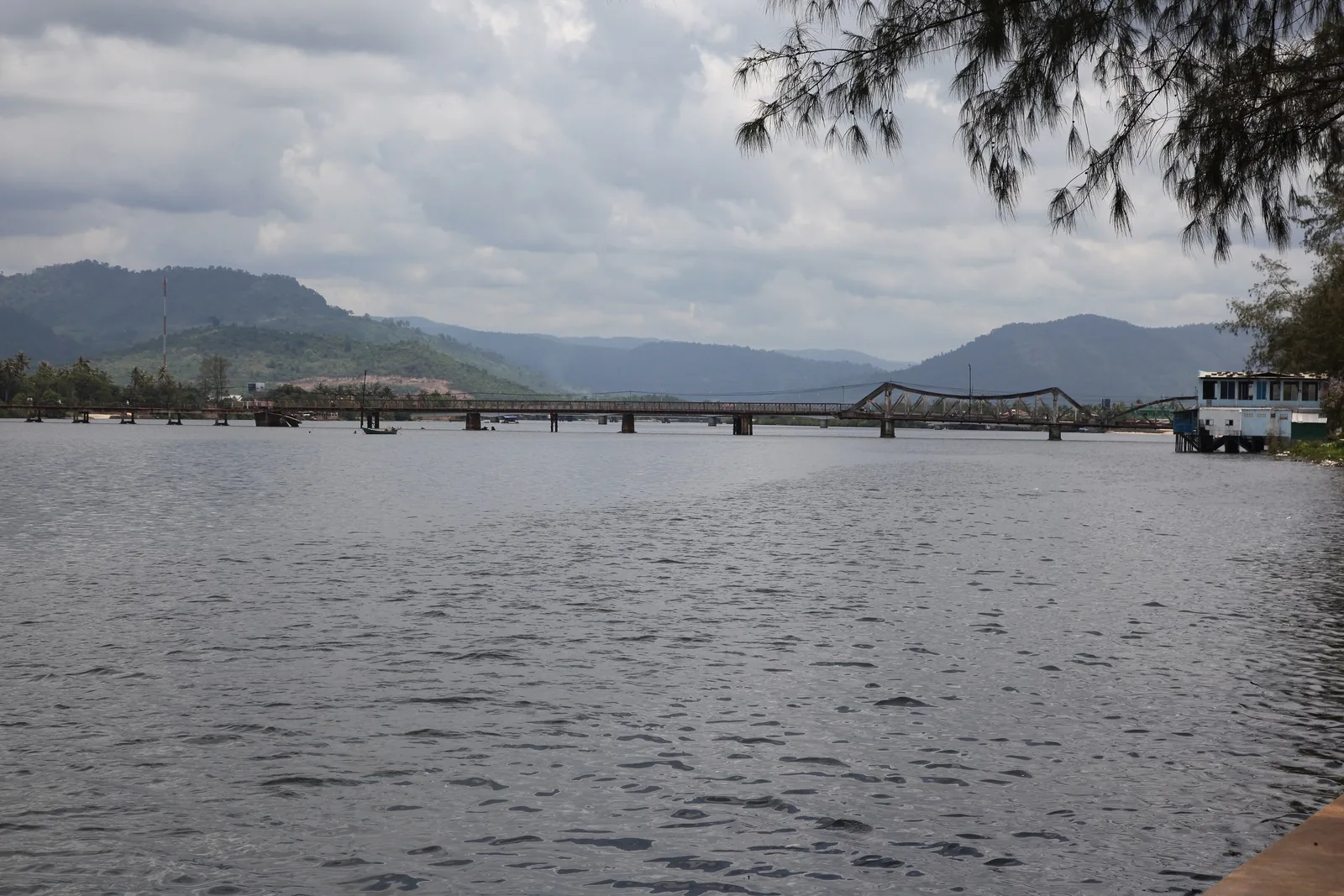 A long bridge spans the wide river in Kampot, backed by mountains under a cloudy sky.