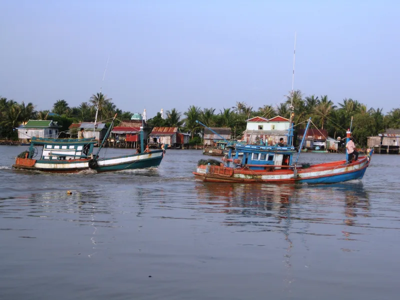 Colorful wooden fishing boats cruise along the Kampot River with a riverside village in the background.