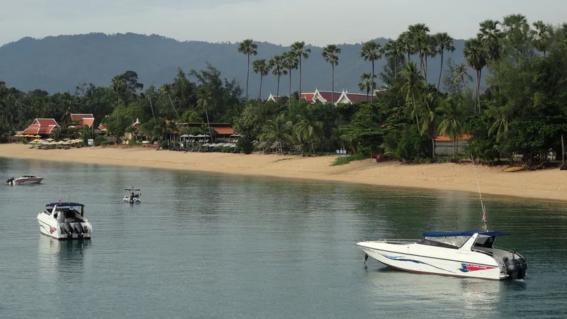 Speedboats float near the calm shore of Maenam Beach on Koh Samui, lined with palm trees and resort roofs.