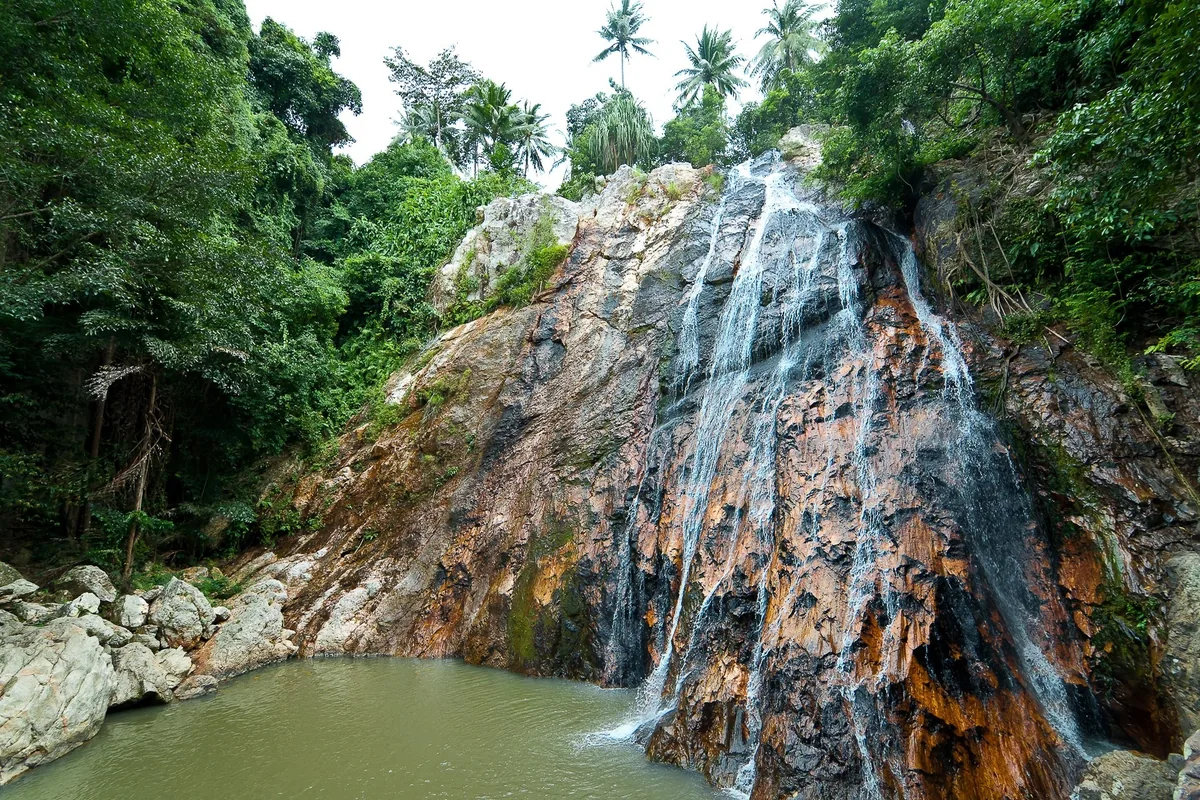Water cascades down a rocky cliff into a pool surrounded by jungle at Na Muang Waterfall in Koh Samui.