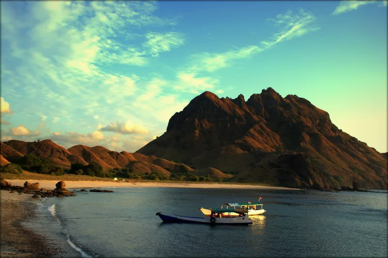 A wooden boat floats near a beach beneath rugged volcanic peaks at Komodo National Park.
