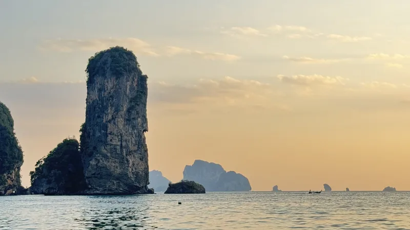 A limestone karst rises from calm sea waters at sunset near Krabi, with distant islands on the horizon.
