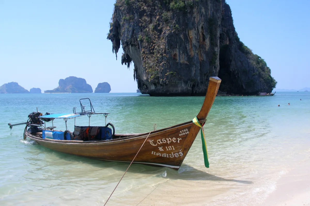 A wooden longtail boat rests on the shore near limestone cliffs at Railay beach in Krabi, Thailand.