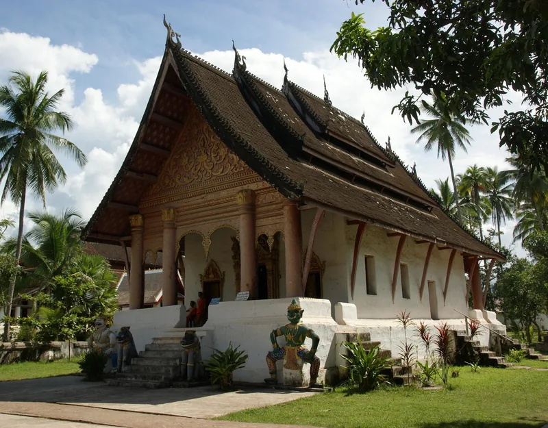 A low sweeping tiered roof and gilded facade mark this Buddhist temple in Luang Prabang, surrounded by palm trees.