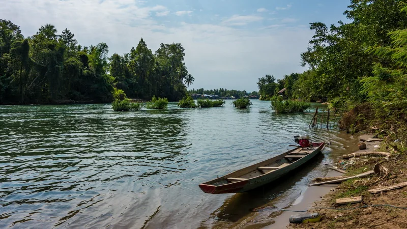 A wooden longtail boat rests on the muddy bank of a river amid the lush greenery of Si Phan Don, Laos.