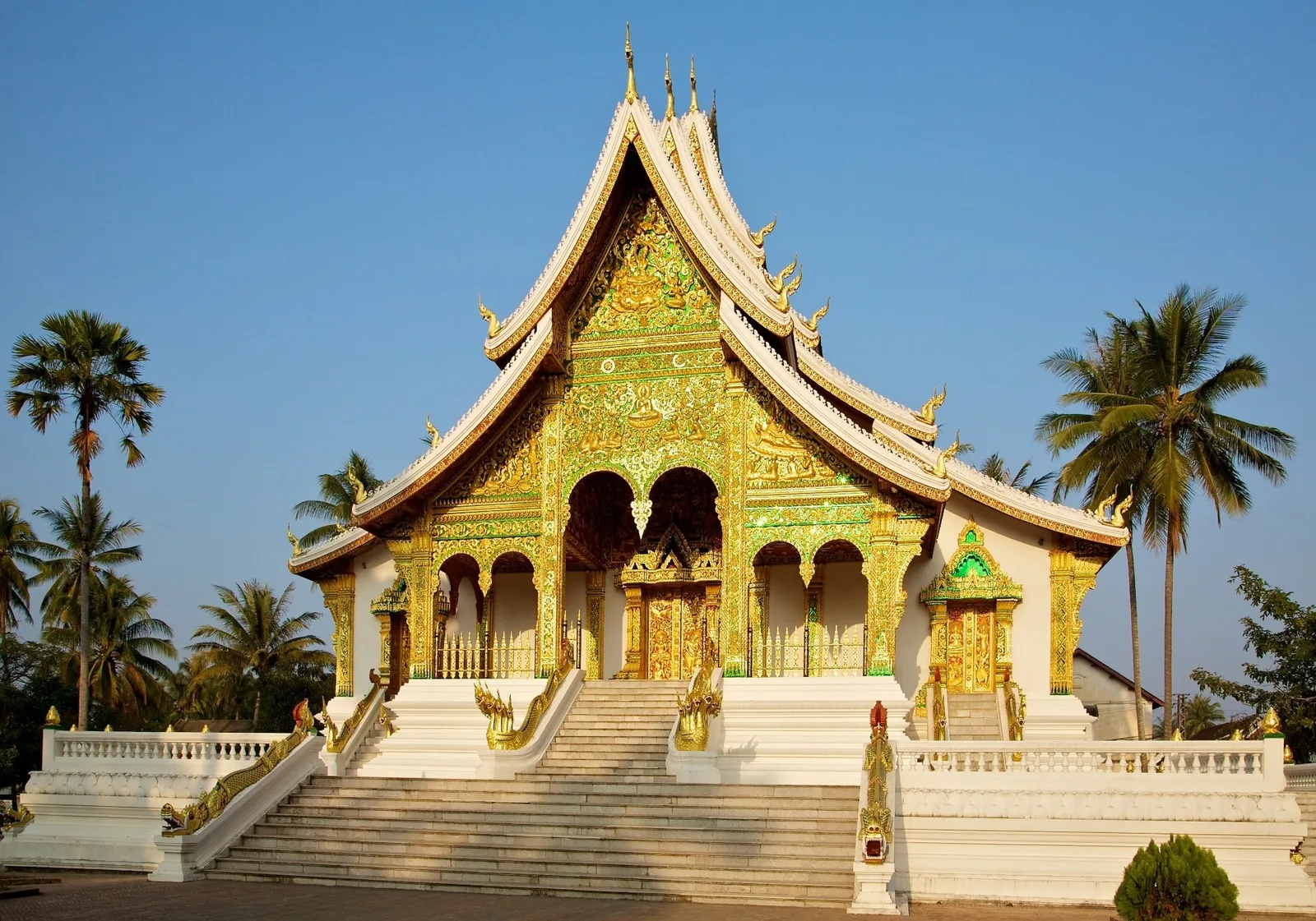 A gilded temple with steep tiered roofs and naga staircases stands among palm trees under a clear sky in Luang Prabang.