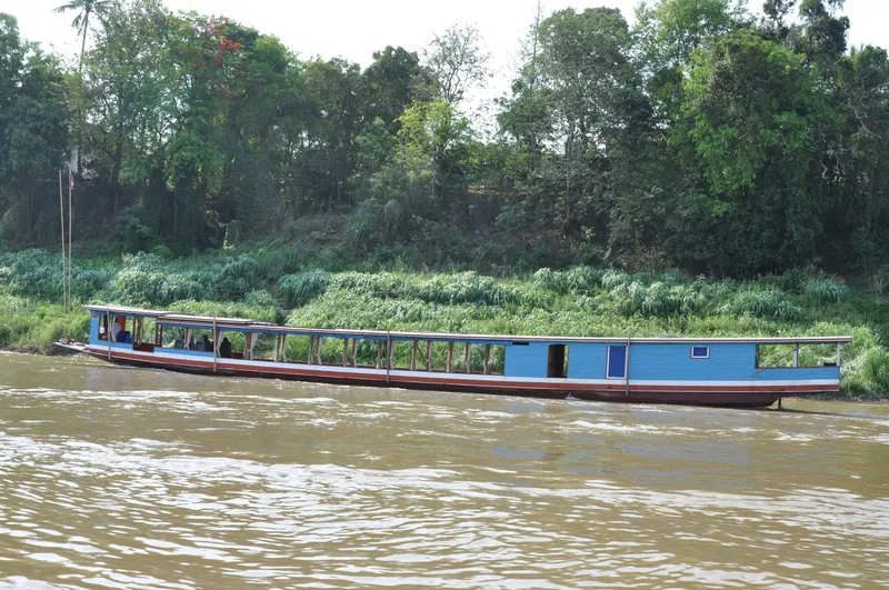 A long blue wooden boat cruises along the muddy Mekong River near lush riverbanks in Luang Prabang.