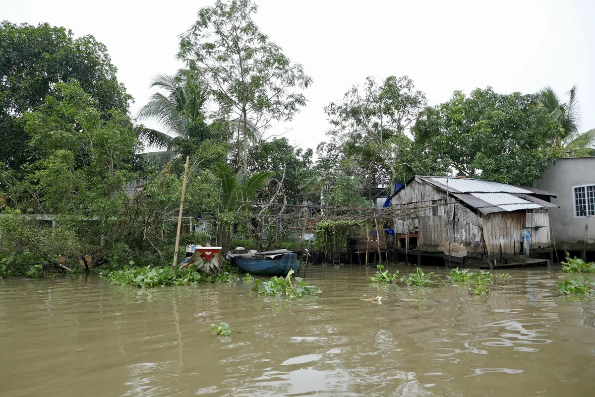 A wooden house on stilts and small boats line the muddy riverbank in the Mekong Delta.