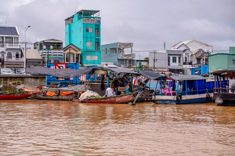 Wooden boats loaded with produce gather on a muddy river in front of colorful buildings at a Mekong Delta floating market.