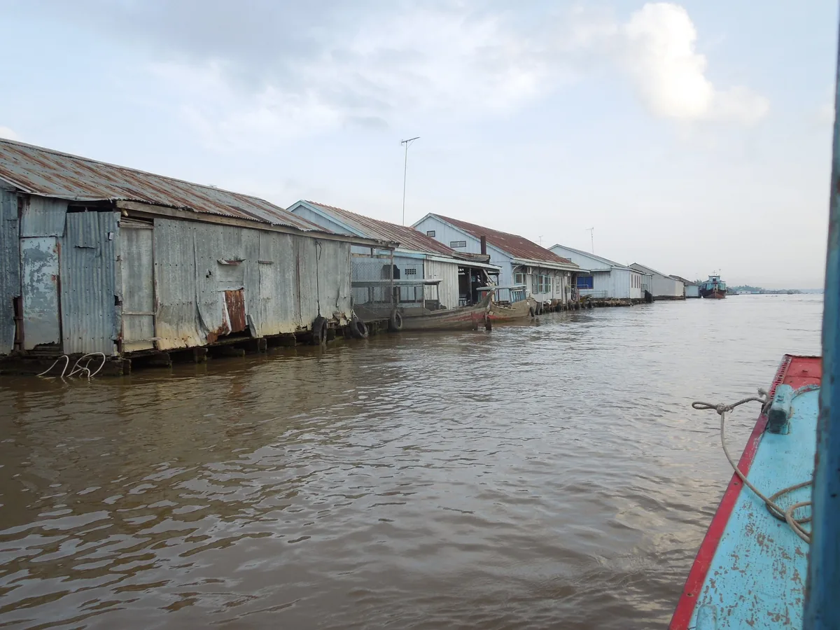 Corrugated metal stilt houses line the muddy river in Chau Doc, Mekong Delta, seen from a boat.