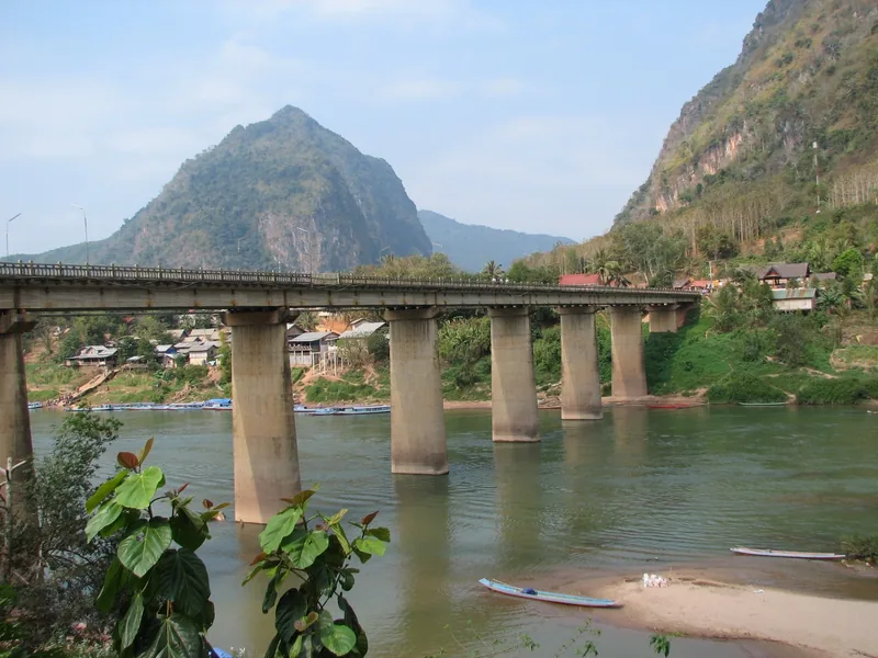 A concrete bridge spans the Nam Ou River at Nong Khiaw, framed by limestone karst mountains and riverside houses.