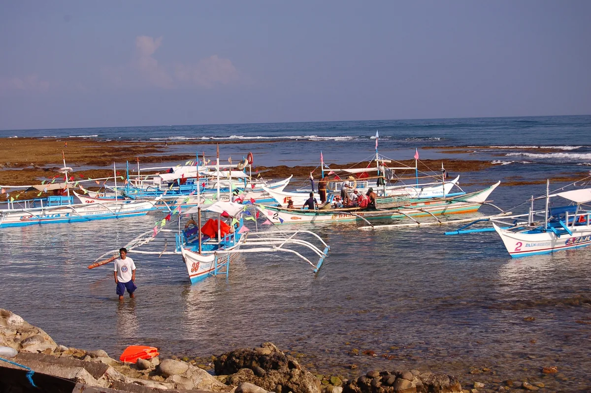 Traditional outrigger boats anchored near a rocky shore in Palawan, with a man wading in shallow water.
