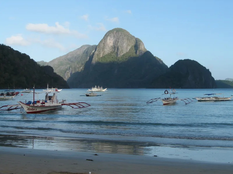 Traditional outrigger boats float on calm water beneath a limestone karst near El Nido, Palawan.