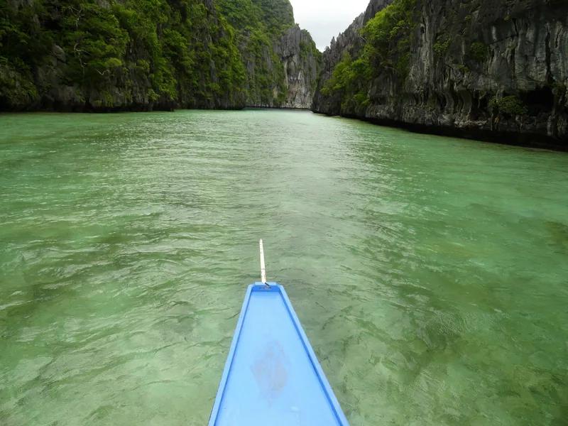 A boat's bow glides through emerald water toward a narrow limestone karst passage in El Nido, Palawan.